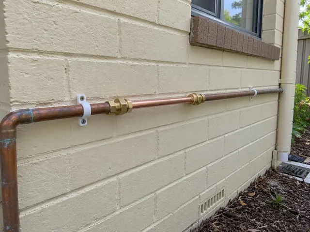 Repaired copper water pipe mounted on a cream brick exterior wall with window and garden bed in Oyster Cove