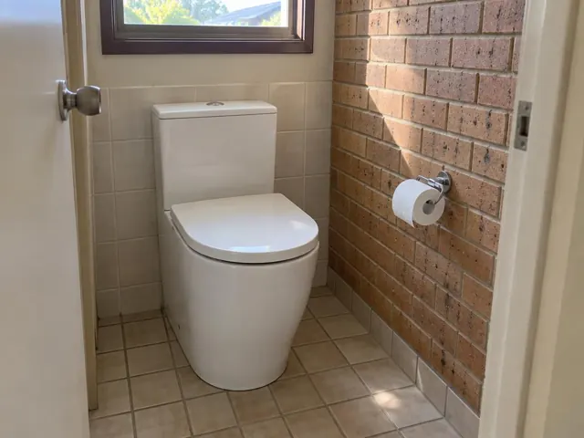 White toilet in a small tiled bathroom with brick feature wall and window in Medowie